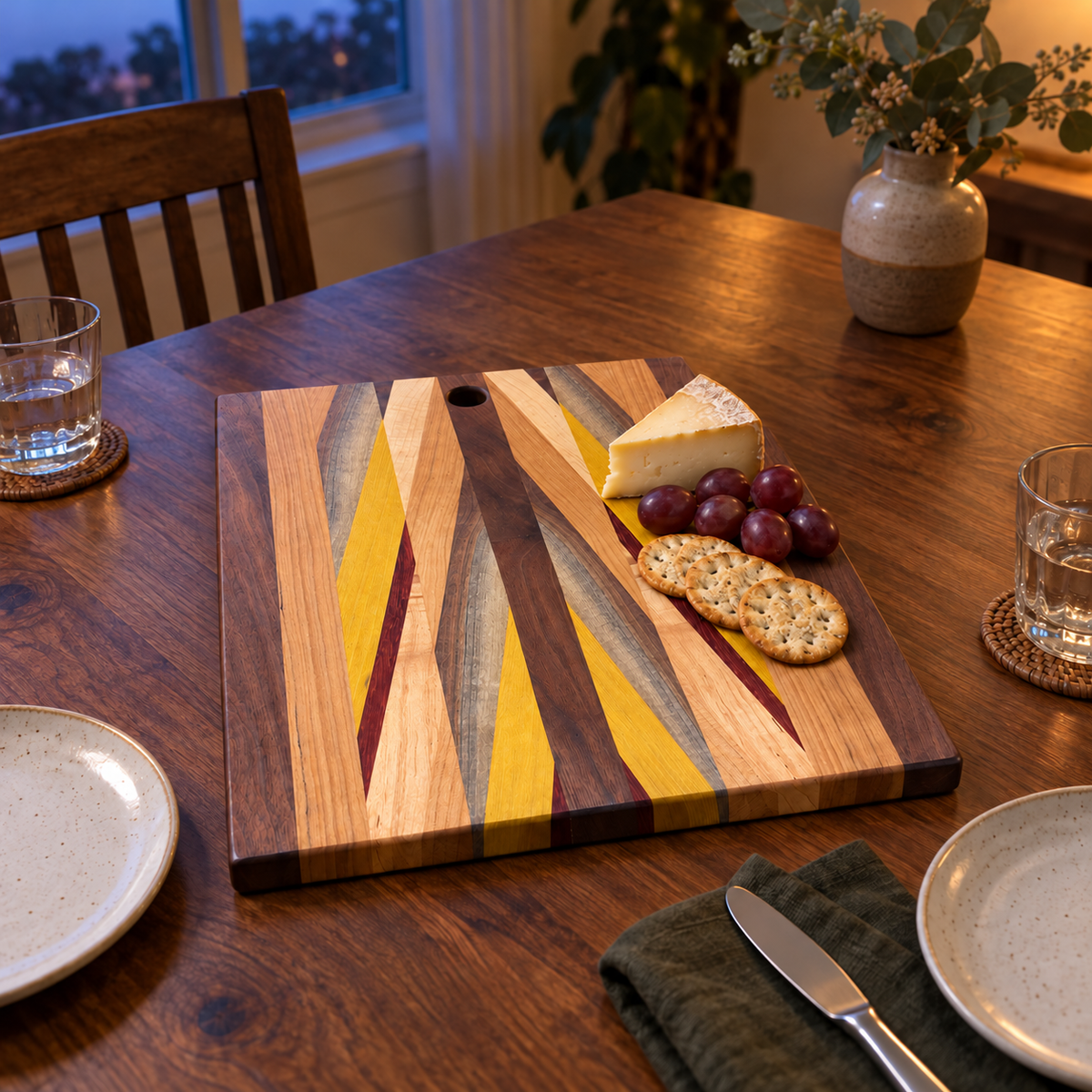 Patterned wood cutting and serving board styled for an evening snack with cheese, grapes and crackers at one end, with two place settings beside it; food and props are not included.