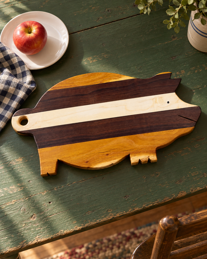 Amish-made pig-shaped wood cutting and serving board with natural cream, brown and golden stripes on a green kitchen table.
