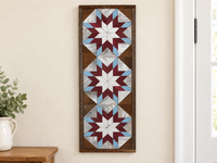 S595 — tall wooden barn quilt banner wall art, three stacked starbursts with white centers, burgundy middles and light blue tips, displayed on an indoor wall in a styled home