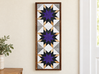 S597 — tall wooden barn quilt banner wall art, three stacked starbursts with purple centers, black middles and natural wood tips, displayed on an indoor wall in a styled home