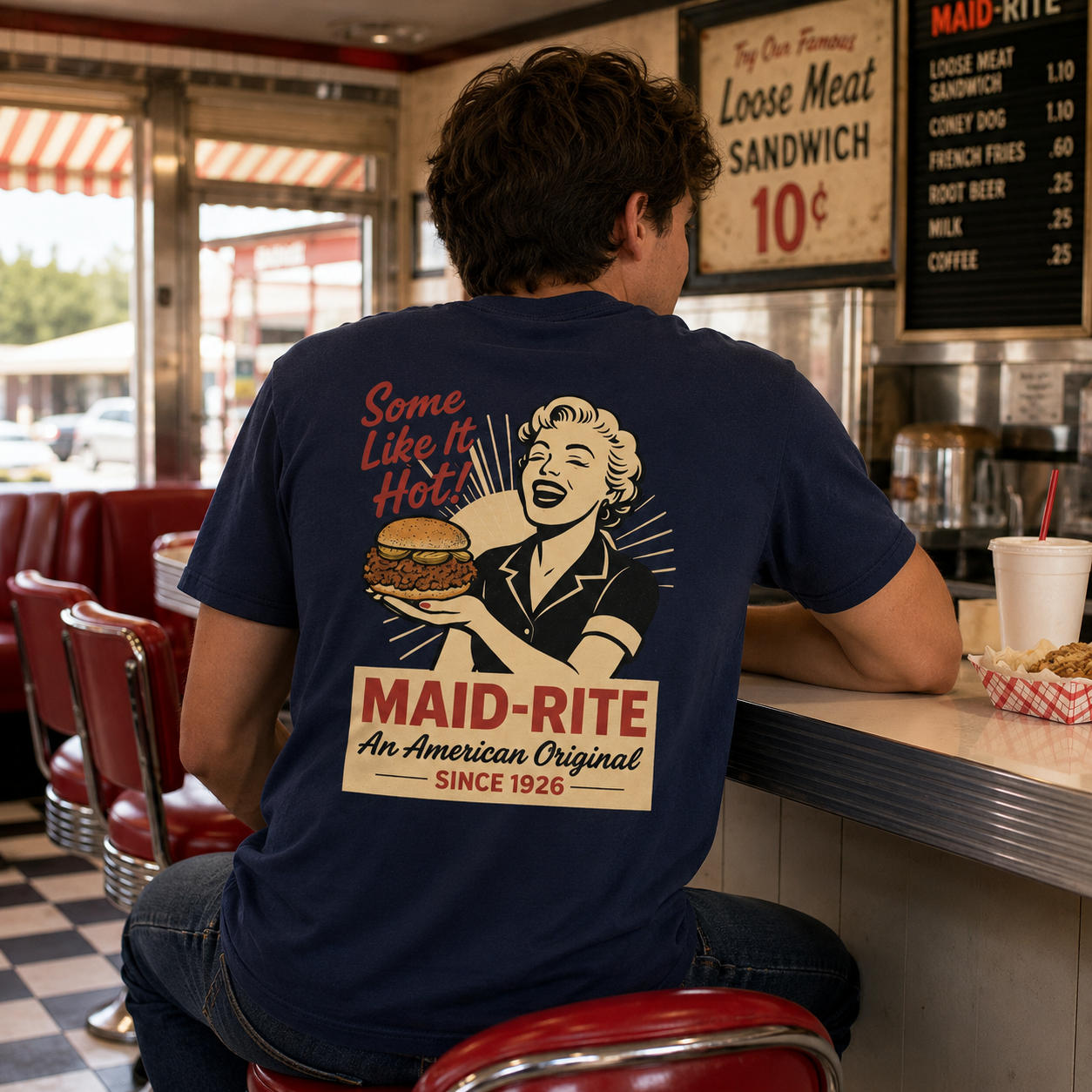 A man sitting at a diner counter wearing a navy blue t-shirt with a retro-style graphic of a woman holding a burger and the text 'Some Like It Hot! MAID-RITE An American Original Since 1926'.